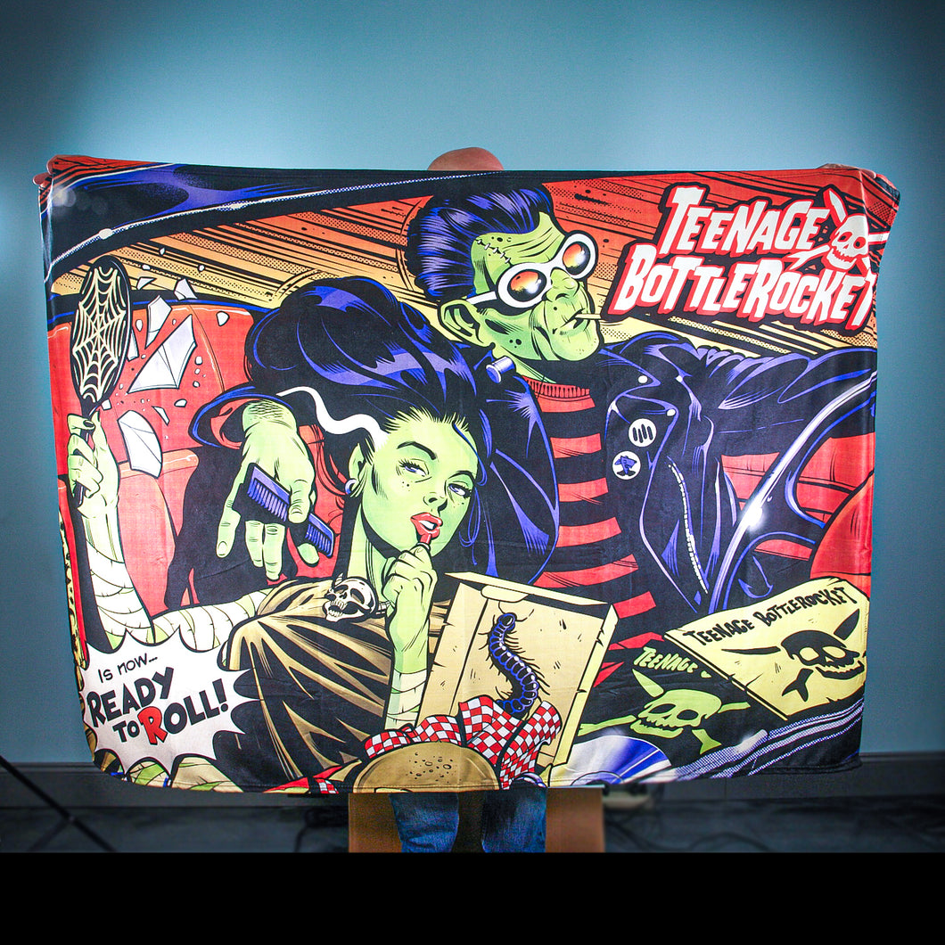 A person holds up a Teenage Bottlerocket blanket with a comic-style design of two green-skinned punks driving, bold patterns, and striking lettering—ideal for fans of the brand.