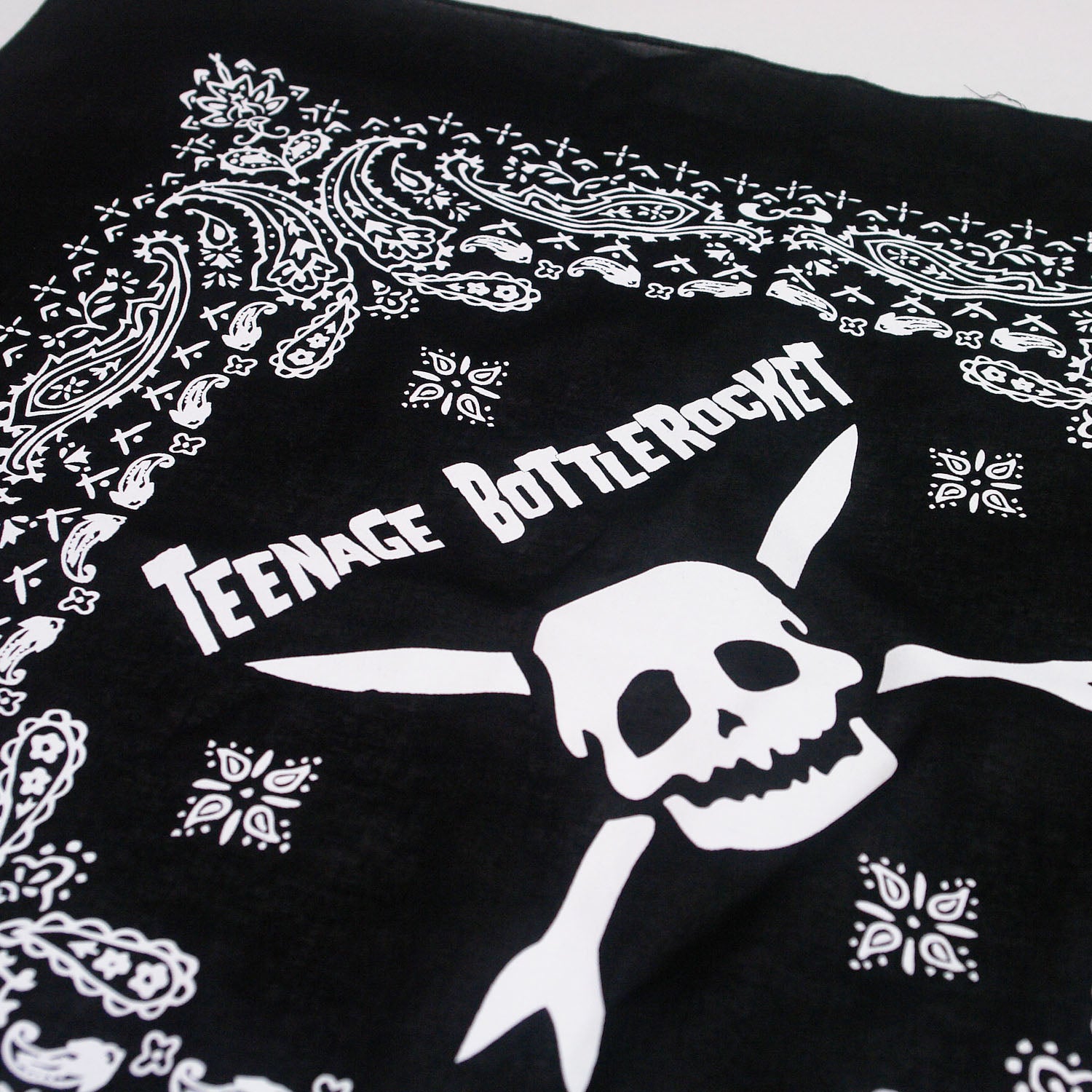 Up close Image of a black bandana against a white background. The bandana says teenage bottlerocket in the center of it in white, and below that is their logo of a white skull head with cross arrows. Surrounding this image is a paisley pattern in white.