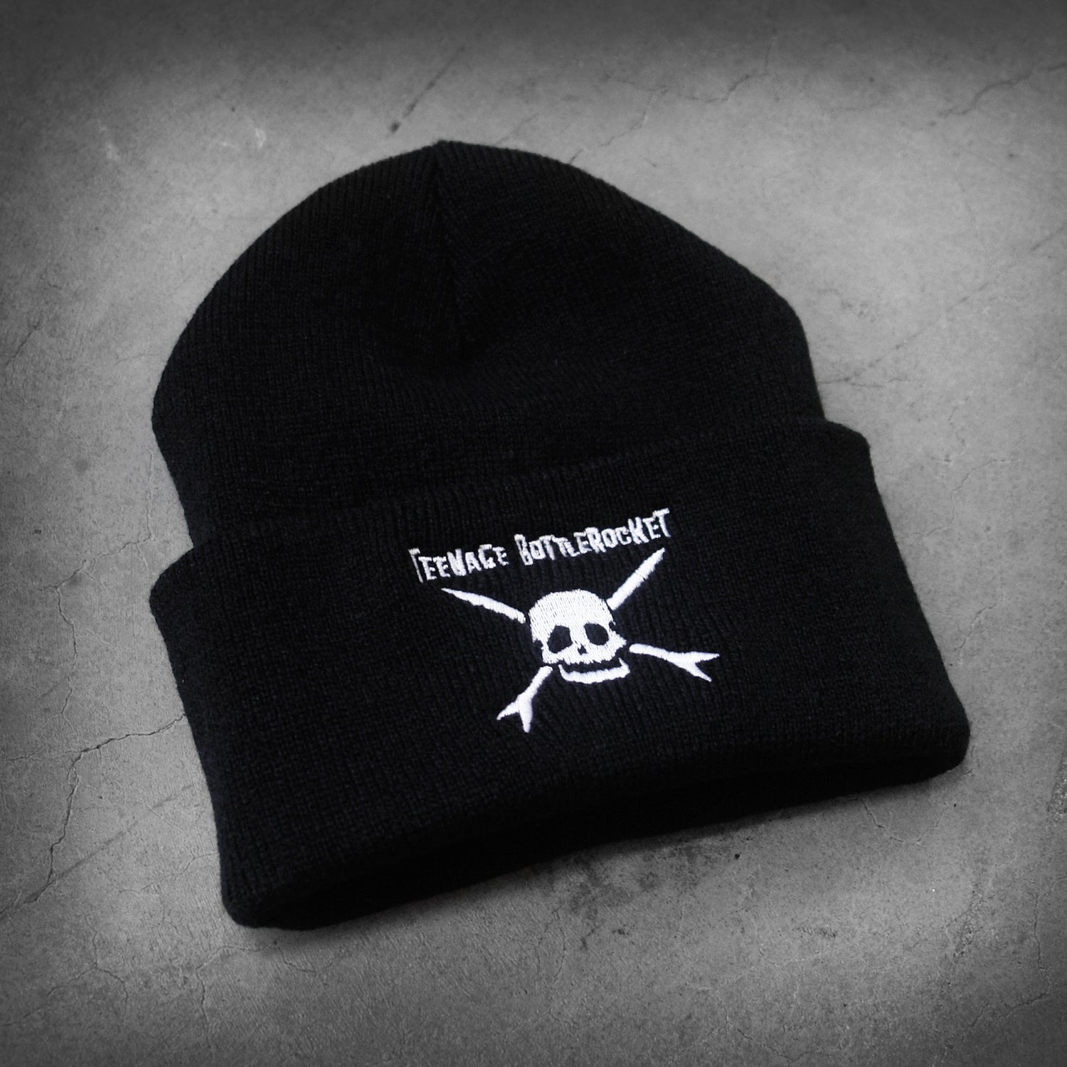 image of a black winter beanie laid flat on a concrete floor. beanie has white embroidery on the front cuff of a skull and teenage bottlerocket above it